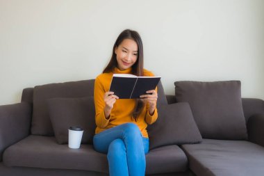 Portre genç Asyalı kadın oturma odasında yastıkla koltukta kitap okudu.