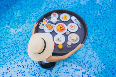 Asyalı güzel bir kadının portresi yüzme havuzunda yüzen kahvaltı tepsisiyle mutlu bir gülümseme