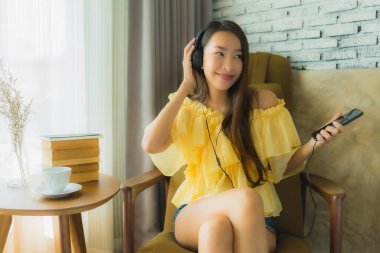 Genç Asyalı bir kadının portresi sandalyeye oturup oturma odasında cep telefonu kahvesi ve kitapla müzik dinlemek.