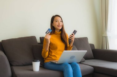 Genç Asyalı kadının oturma odasında online alışveriş yapmak için akıllı cep telefonu ve kredi kartı olan dizüstü bilgisayarı kullandığı portre.