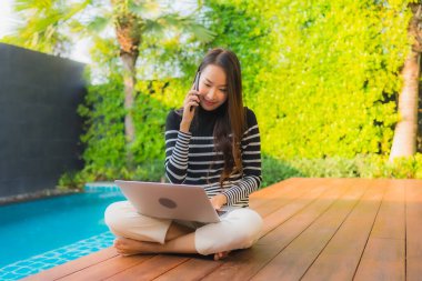 Asyalı genç bir kadının otel havuzunda akıllı cep telefonlu dizüstü bilgisayar kullanarak yüzdüğü portre.