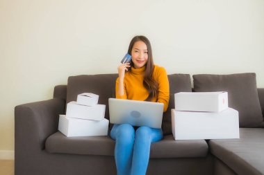 Asyalı genç bir kadının portresi, oturma odasındaki koltukta paket kutusuyla çalışmak için dizüstü bilgisayar kullanıyor.