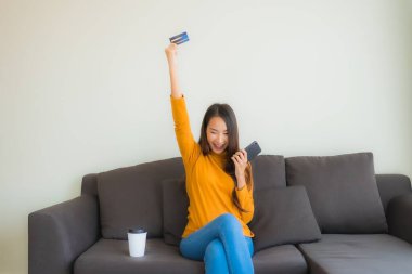 Genç Asyalı kadının oturma odasında online alışveriş yapmak için akıllı cep telefonu ve kredi kartı olan dizüstü bilgisayarı kullandığı portre.