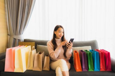 Portre güzel genç Asyalı kadın dizüstü bilgisayar ya da akıllı cep telefonu kullanarak oturma odasındaki alışveriş çantasının etrafındaki kanepede alışveriş yapıyor.