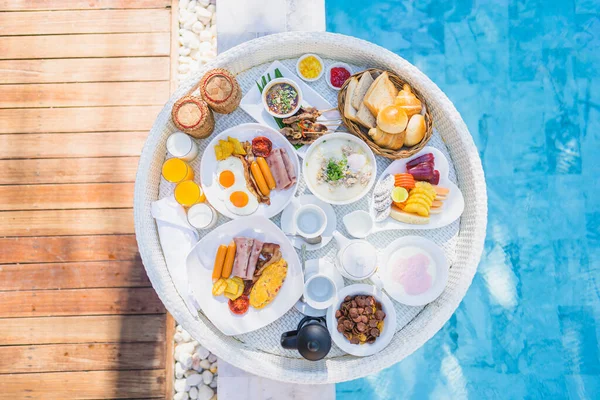 Floating breakfast set in tray with fried egg omelette sausage ham bread fruit milk juice coffee and other around swimming pool