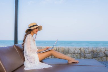 Plaj denizi çevresinde çalışmak için dizüstü bilgisayarı kullanan Asyalı genç bir kadının portresi.