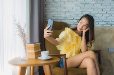 Genç Asyalı kadın kahve fincanı ile cep telefonu kullanıyor ve kitap okuyor oturma odasındaki sandalyeye oturuyor.