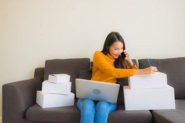 Asyalı genç bir kadının portresi, oturma odasındaki koltukta paket kutusuyla çalışmak için dizüstü bilgisayar kullanıyor.