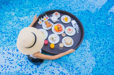 Asyalı güzel bir kadının portresi yüzme havuzunda yüzen kahvaltı tepsisiyle mutlu bir gülümseme