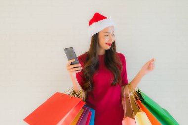 Güzel Asyalı genç kadınların portresi Noel Baba şapkası ve alışveriş çantası, akıllı cep telefonu ve kredi kartı takıyor.