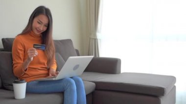 Genç Asyalı kadının oturma odasında online alışveriş yapmak için akıllı cep telefonu ve kredi kartı olan dizüstü bilgisayarı kullandığı portre.