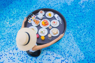 Asyalı güzel bir kadının portresi yüzme havuzunda yüzen kahvaltı tepsisiyle mutlu bir gülümseme