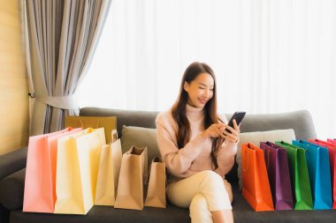 Portre güzel genç Asyalı kadın dizüstü bilgisayar ya da akıllı cep telefonu kullanarak oturma odasındaki alışveriş çantasının etrafındaki kanepede alışveriş yapıyor.
