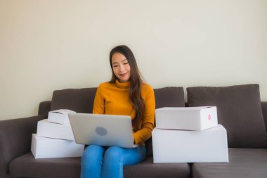 Asyalı genç bir kadının portresi, oturma odasındaki koltukta paket kutusuyla çalışmak için dizüstü bilgisayar kullanıyor.