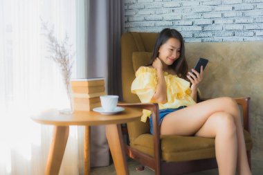 Genç Asyalı kadın kahve fincanı ile cep telefonu kullanıyor ve kitap okuyor oturma odasındaki sandalyeye oturuyor.