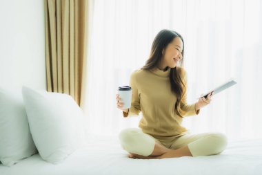 Genç Asyalı kadın, elinde kahve fincanıyla yatak odasında kitap okuyor.