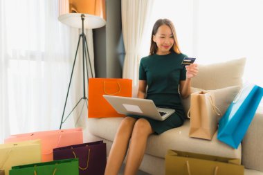 Güzel Asyalı genç kadınların portresi kanepeye oturup bilgisayar bilgisayarı ya da akıllı ve cep telefonuyla online alışveriş için kredi kartı kullanırlar.