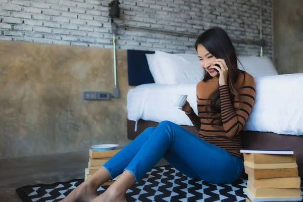Young asian woman using smart mobile phone with read book and coffee cup in bedroom interior