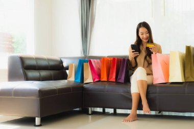 Güzel Asyalı bir kadının portresi. İnternetten alışveriş yapıyor. Kredi kartı ve akıllı telefonuyla oturma odasındaki alışveriş çantasının etrafında.