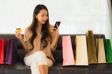 Güzel Asyalı bir kadının portresi. İnternetten alışveriş yapıyor. Kredi kartı ve akıllı telefonuyla oturma odasındaki alışveriş çantasının etrafında.