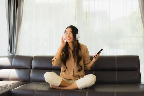 ポートレートの美しい若いアジアの女性は 音楽を聴くためのヘッドフォンとソファの上にスマートフォンや携帯電話を使用 — ストック写真