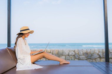 Plaj denizi çevresinde çalışmak için dizüstü bilgisayarı kullanan Asyalı genç bir kadının portresi.