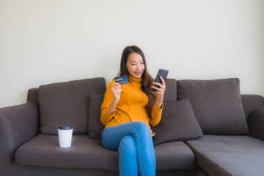 Genç Asyalı kadının oturma odasında online alışveriş yapmak için akıllı cep telefonu ve kredi kartı olan dizüstü bilgisayarı kullandığı portre.