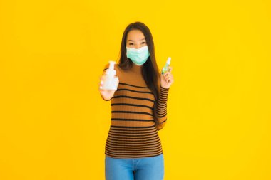 Koronavirüsten korunmak için elinde maske spreyi ve jel şişesi olan güzel Asyalı kadın portresi ya da sarı izole edilmiş arka planda kovid19.
