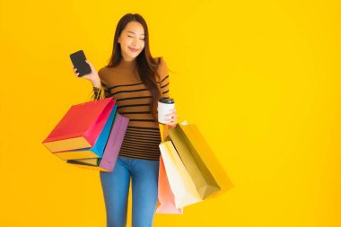 Güzel, genç Asyalı bir kadın sarı izole arka planda kahve fincanı ve renkli alışveriş çantasıyla akıllı cep telefonu veya cep telefonu kullanır.