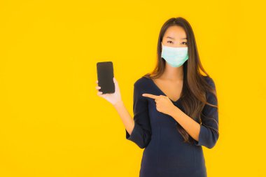 Maskeli ve akıllı cep telefonlu güzel bir Asyalı kadın portresi veya izole edilmiş sarı arka planda cep telefonu.