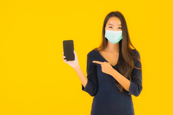 亚洲年轻貌美的女人 戴着面具 带着智能手机或手机 背景是黄色独立的 — 图库照片