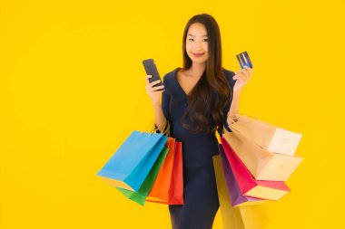 Portre güzel genç Asyalı kadın alışveriş çantası kredi kartı ve akıllı cep telefonu sarısı izole bir arka plan.