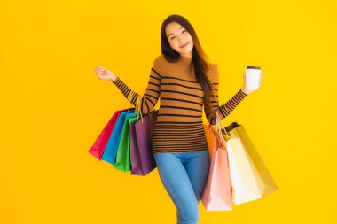 Güzel, genç Asyalı bir kadın sarı izole arka planda kahve fincanı ve renkli alışveriş çantasıyla akıllı cep telefonu veya cep telefonu kullanır.