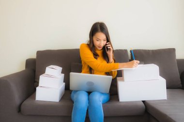 Asyalı genç bir kadının portresi, oturma odasındaki koltukta paket kutusuyla çalışmak için dizüstü bilgisayar kullanıyor.