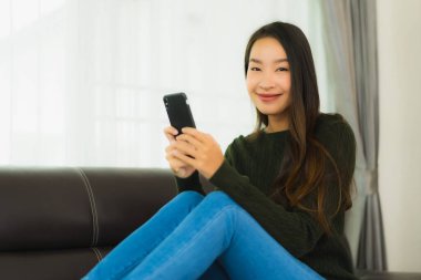 Güzel bir Asyalı kadın oturma odasındaki koltukta akıllı cep telefonu ya da cep telefonu kullanıyor.