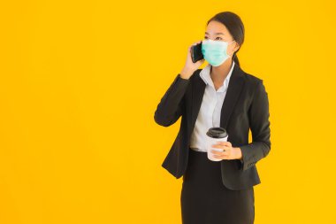 Portre güzel genç Asyalı kadın maske takıyor. Cep telefonu ve kahve ile covid19 ve koronavirüs konseptini sarı izole arka planda kullanın.