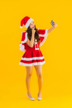 Güzel bir Asyalı kadın portresi Noel kıyafetleri giyer ve sarı izole edilmiş arka planda akıllı cep telefonu kullanır.