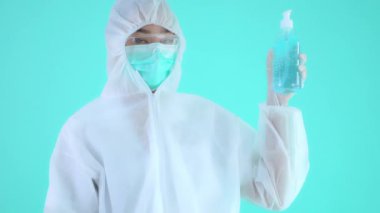 Korumalı tıbbi giysi ve maske takan Asyalı bir kadının turkuazda izole edilmiş bir dezenfektan şişesi görüntüsü.