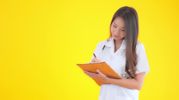 黄色の上に隔離されたクリップボードに書いている美しいアジアの医師の女性の映像 — ストック動画