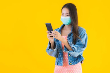 Güzel Asyalı genç bir kadının portresi kovid19 ya da koronavirüsü korumak için maske takar. Sarı izole edilmiş arka planda akıllı cep telefonu kullanın.