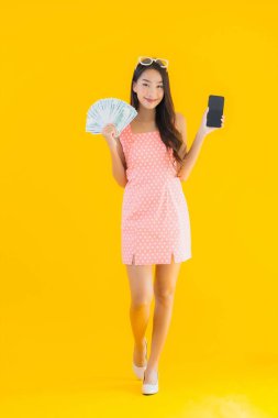 Güzel bir Asyalı kadının portresi, sarı izole edilmiş arka planda cep telefonuyla yüklü miktarda nakit ve para gösteriyor.
