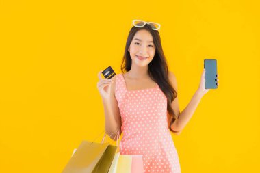 Renkli alışveriş çantalı güzel Asyalı kadın akıllı cep telefonu ve sarı izole edilmiş arka planda kredi kartı.