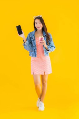 Güzel bir Asyalı kadın portresi telefon patlamış mısırı ve sarı izole edilmiş bir arka planda film izlemekten memnun.