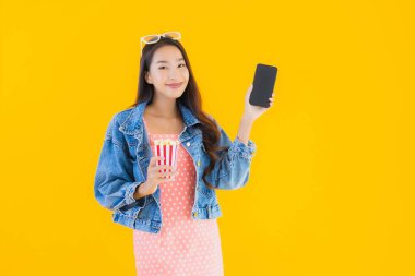 Güzel bir Asyalı kadın portresi telefon patlamış mısırı ve sarı izole edilmiş bir arka planda film izlemekten memnun.