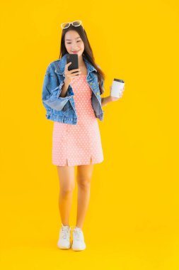 Kahve fincanı ve sarı izole edilmiş arka planda akıllı cep telefonlu güzel Asyalı kadın portresi.