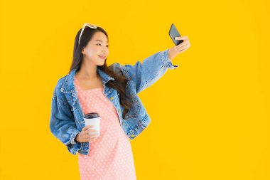 Kahve fincanı ve sarı izole edilmiş arka planda akıllı cep telefonlu güzel Asyalı kadın portresi.