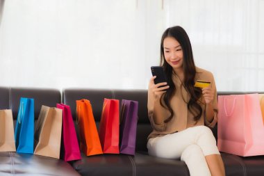 Güzel Asyalı bir kadının portresi. İnternetten alışveriş yapıyor. Kredi kartı ve akıllı telefonuyla oturma odasındaki alışveriş çantasının etrafında.