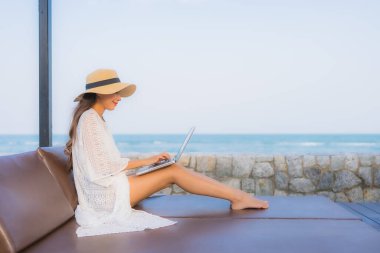 Plaj denizi çevresinde çalışmak için dizüstü bilgisayarı kullanan Asyalı genç bir kadının portresi.