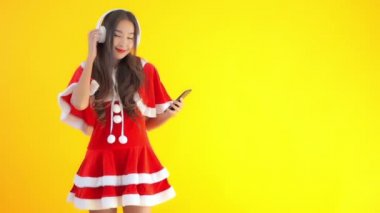 Noel Baba kostümlü güzel bir Asyalı kadının, sarı renkte izole edilmiş akıllı telefon ve kulaklıklarla müzik dinlediği görüntüler.