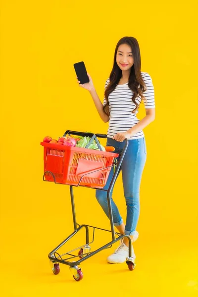 在超级市场和购物车上用黄色独立背景的智能手机购物的漂亮年轻亚洲女人的画像 — 图库照片